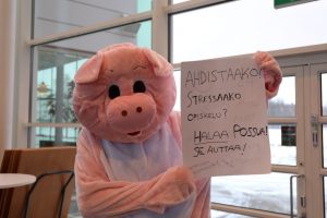 Possuhahmo kädessään kyltti, jossa lukee: "Ahdistaako, stressaako opiskelu? Halaa possua! Se auttaa!"
