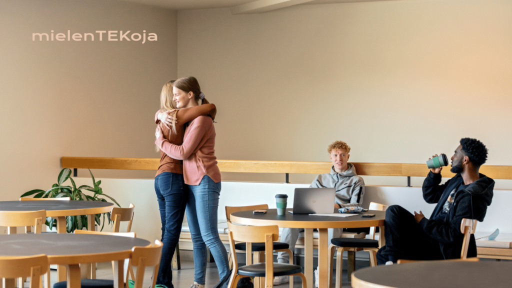 Kaksi naisoletettua halaavat valoisassa huoneessa, jossa istuu kaksi miesoletettua