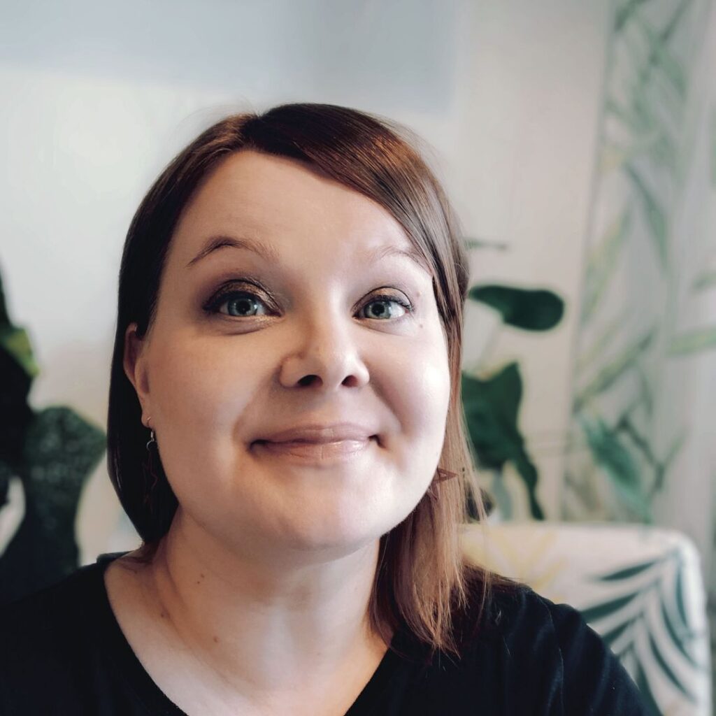 Smiling woman who has a light red bob and black shirt. In the background there's a chair with leaf patterns and house plants.