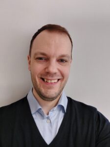 Hymyilevä miesoletettu vaaleaa seinää vasten. Henkilöllä on tummat lyhyet hiukset ja tumma parransänki. Päällään Tommilla on vaalea kaulupaita ja tumma neule