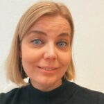 Vaalea, polkkatukkainen naisoletettu, jolla siniset silmät ja voimakkaat kulmakarvat sekä korkeat poskipäät. Henkilö hymyilee ja hänellä on yllään tumma kilpikonnakauluksella varustettu pusero.