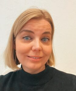 Vaalea, polkkatukkainen naisoletettu, jolla siniset silmät ja voimakkaat kulmakarvat sekä korkeat poskipäät. Henkilö hymyilee ja hänellä on yllään tumma kilpikonnakauluksella varustettu pusero.