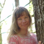 En kvinnlig förmodad person bredvid stammen på ett tjockt lövträd. Hon har ganska blont långt hår och ljus hy. Personen ler lite. Hon är klädd i en rosa mönstrad skjorta. I bakgrunden syns trädens sommargrönska.