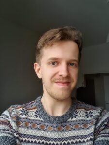 Miesoletettu henkilö, jolla keskiruskeat lyhyet hiukset ja viikset. Henrillä on yllään kuviollinen harmaa neule