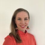 Naisoletettu henkilö, jolla on pitkät vaaleat hiukset. Hiukset laskeutuva Leenan oikealle olkapäälle. Leena hymyilee ja hänellä on isot oranssiset korvakorut sekä oranssinen paita, jossa kilpikonnakaulus.