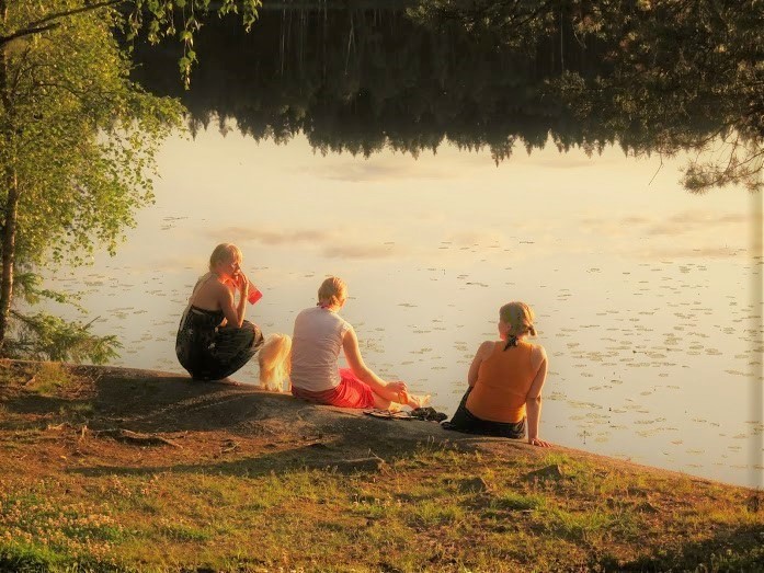 Kolme naisoletettua istuu kalliolla selin, taustallaan järvi, aurinko paistaa. Kahden naisen välistä näkyy valkoinen koiran häntä.