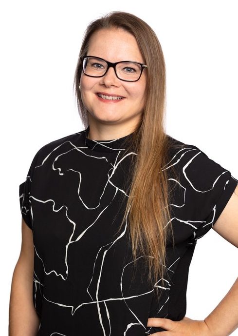 Brown-haired smiling woman with dark-rimmed glasses. The woman is wearing a black t-shirt with white stripes.