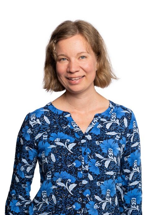 A smiling woman with quite short, blond hair. She is wearing a flower patterned, blue-toned blouse.