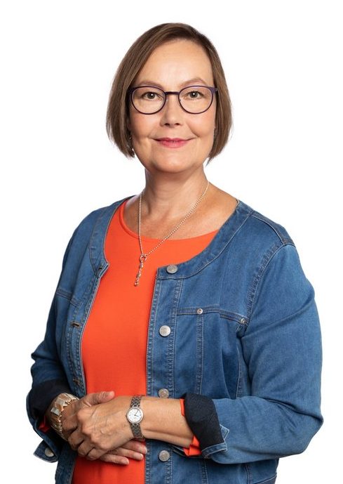 Brown-haired smiling woman with glasses. The woman is wearing an orange dress with a denim jacket on top.