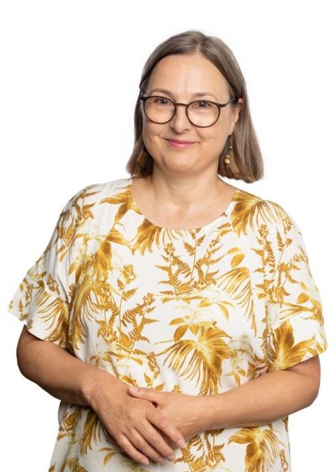 A woman smiles and looks at the camera. She has light brown, semi-long hair and glasses. She is wearing a short-sleeved yellow-white pattern dress.