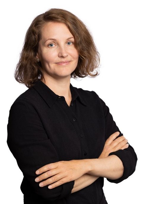 A female presumed person in front of a light background. The person has brown shoulder-length hair and wears a black shirt dress. The person is smiling and looking at the camera.