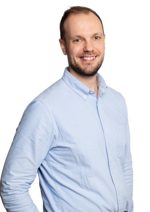 A male presumed person in front of a white background. The person has dark short hair. The person smiles and looks straight into the camera. He is wearing a light blue shirt.