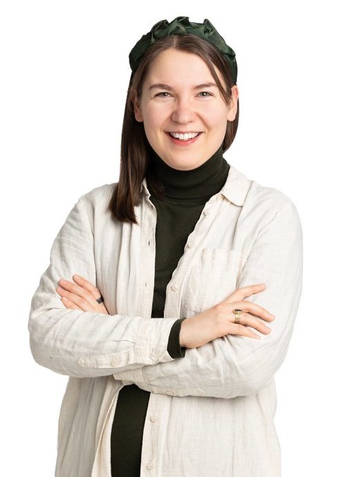 Smiling woman with long brown hair. The woman is wearing a light dress with a dark green polo shirt underneath.