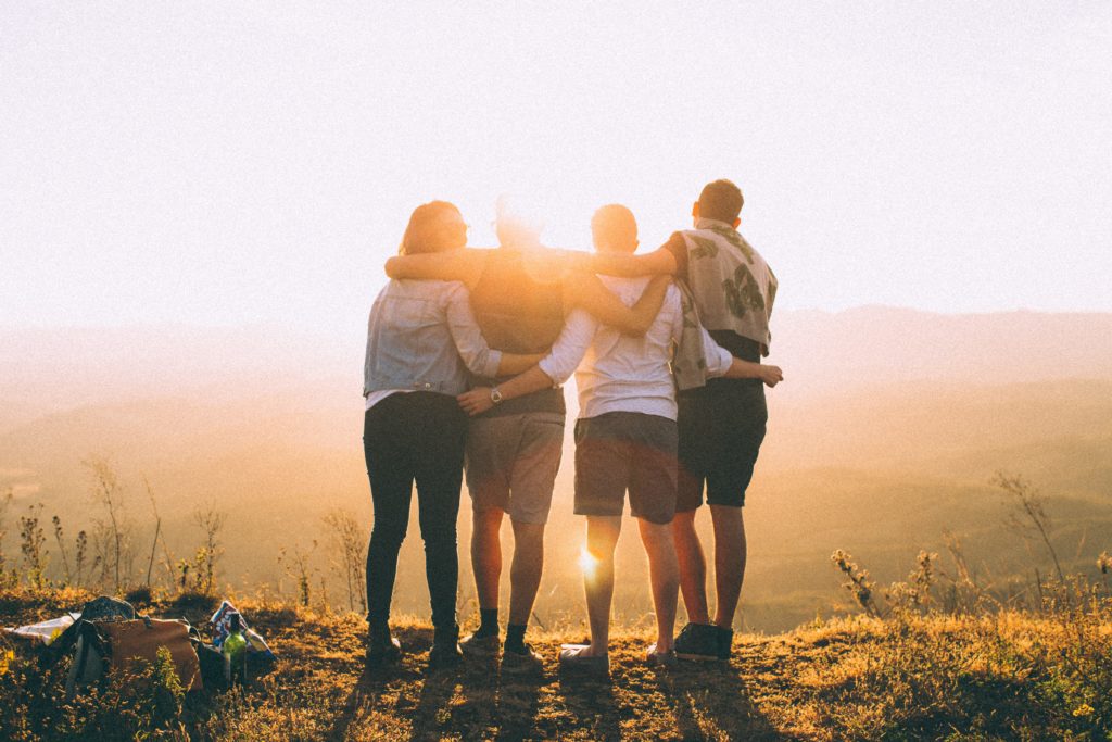 Kuvassa neljä nuorta ihmistä seisomassa rinnakkain kädet toistensa harteilla katselemassa korkealla paikalle auringonlaskun suuntaan, kesävaatteissa, kuva otettu takaapäin.